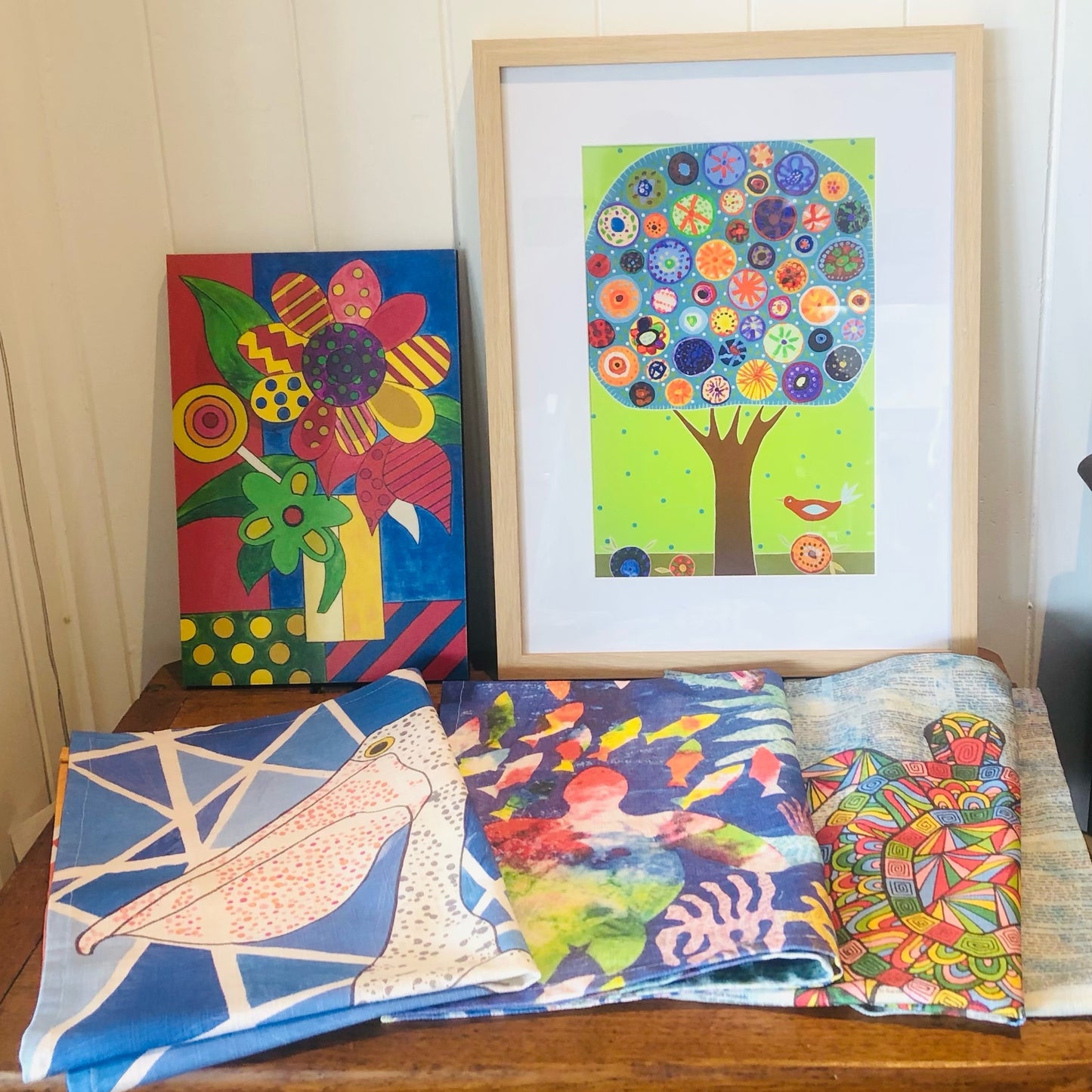 Custom-made fundraising tea towels folded at the front of timber table with two artworks leaning agains the wall.